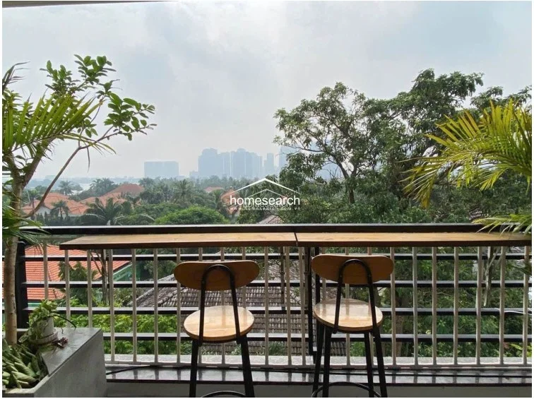 This balcony serves as a scenic outdoor retreat, featuring a built-in bar-style counter and two stools that overlook a lush green landscape and distant city skyline. The space is well-maintained, incorporating potted plants and a metal railing, making it an ideal spot for relaxation.