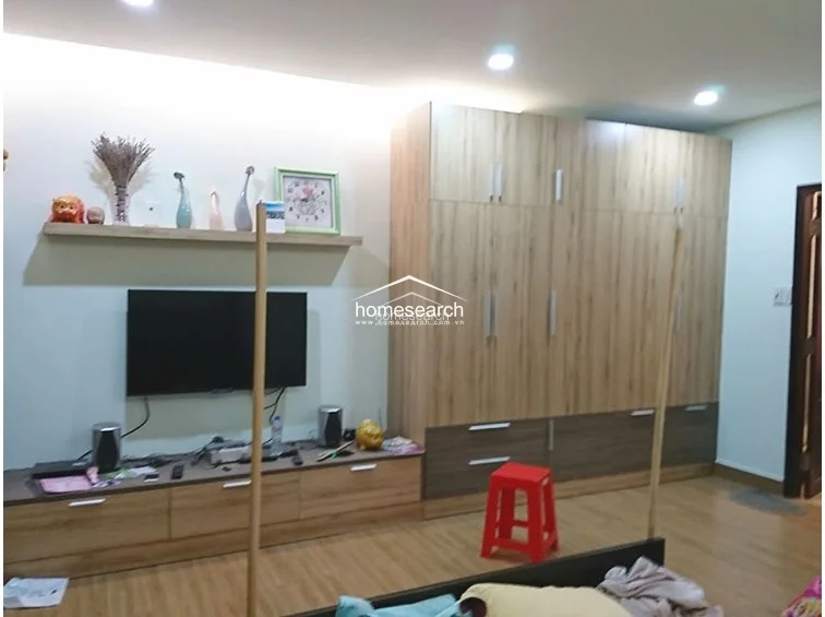 This bedroom view highlights a large, floor-to-ceiling wooden wardrobe that provides extensive storage space. The room includes a wall-mounted TV, a floating display shelf, and wood-look flooring, maintaining a consistent, well-maintained aesthetic.