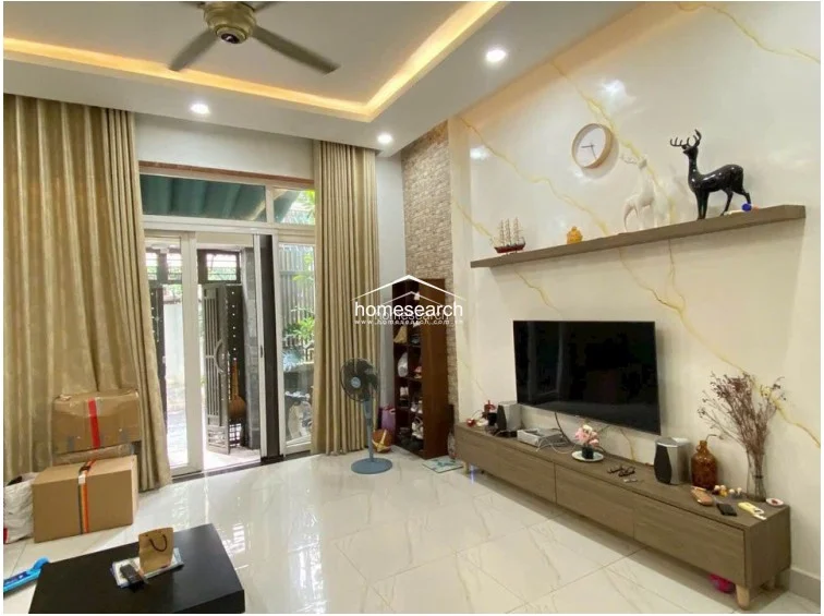 This living room features a bright, modern aesthetic with glossy white floor tiles and a feature wall adorned with decorative gold-veined paneling. A large glass door with floor-to-ceiling beige curtains provides access to an outdoor area, allowing for ample natural light. The room is furnished with a sleek, low-profile wooden media console and recessed ceiling lighting.