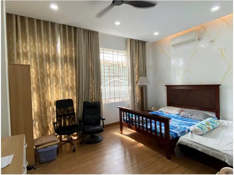 This bedroom features a warm, neutral color scheme with wood-look flooring and a distinctive marble-patterned accent wall. The room is equipped with a ceiling fan, a wall-mounted AC unit, and floor-to-ceiling curtains that frame a window with security bars.
