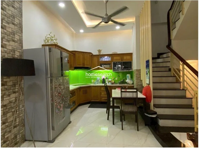 This open-concept kitchen and dining area features warm wooden cabinetry, a vibrant green glass backsplash, and sleek white tile flooring. The space is efficiently laid out with a central dining table and a staircase with a wooden handrail, leading to upper levels.