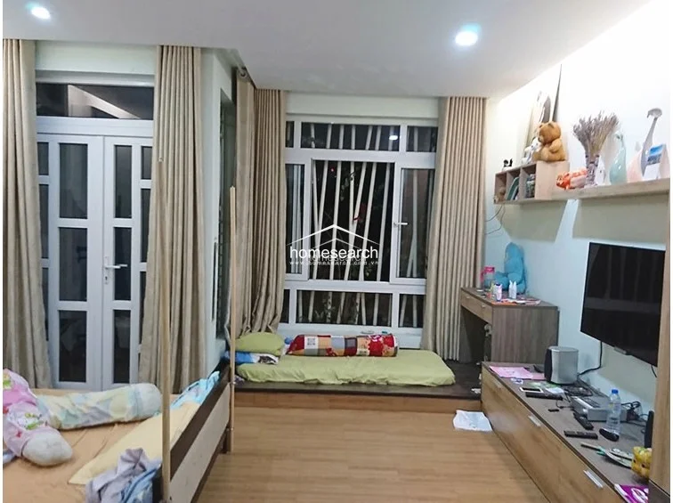 This bedroom layout includes a direct access point to an outdoor balcony through a glass-paned door, enhancing the room's flow and natural light. The space is furnished with a bed, a desk, and a wall-mounted TV, all set against a neutral backdrop with light-colored walls and wood-look flooring.
