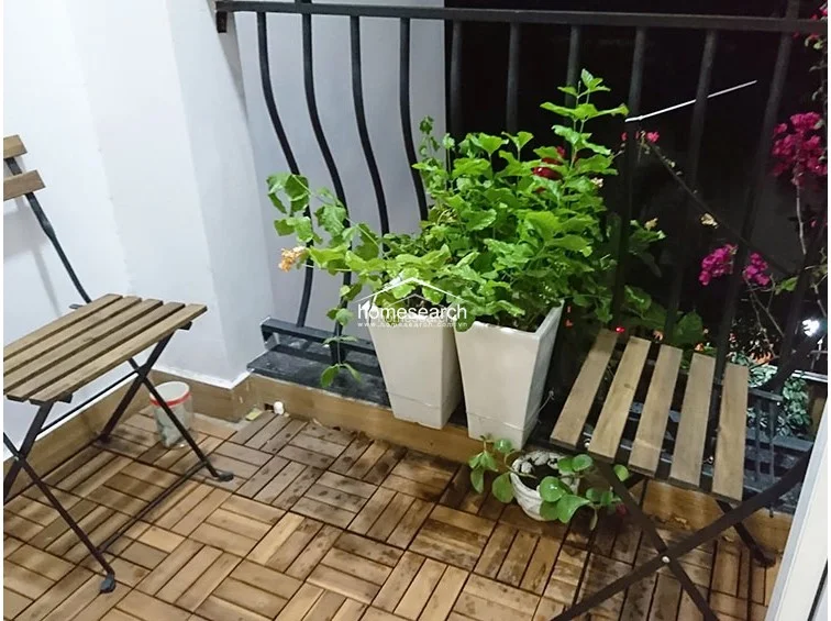 This small, private balcony features durable wood-style deck tiles and a black metal railing, creating a cozy outdoor retreat. It is furnished with two folding wooden chairs and potted plants, offering a modest but well-utilized space for relaxation.
