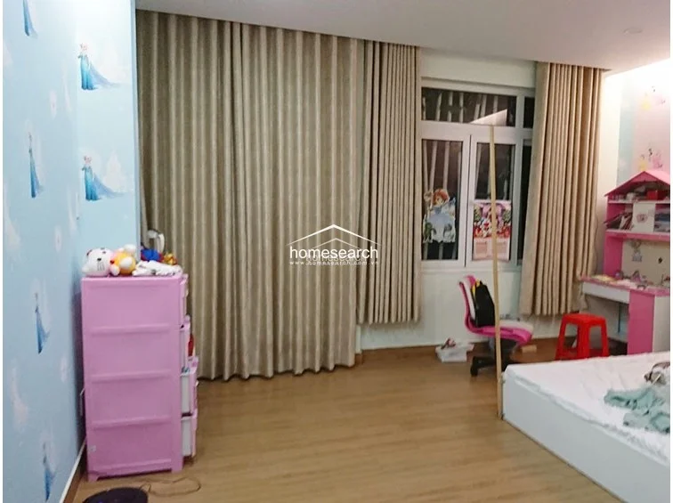 This bedroom is styled as a children's room, featuring light-colored wood flooring and floor-to-ceiling curtains that provide ample privacy. The space is furnished with a pink storage unit and a matching study desk, creating a bright and well-organized environment.