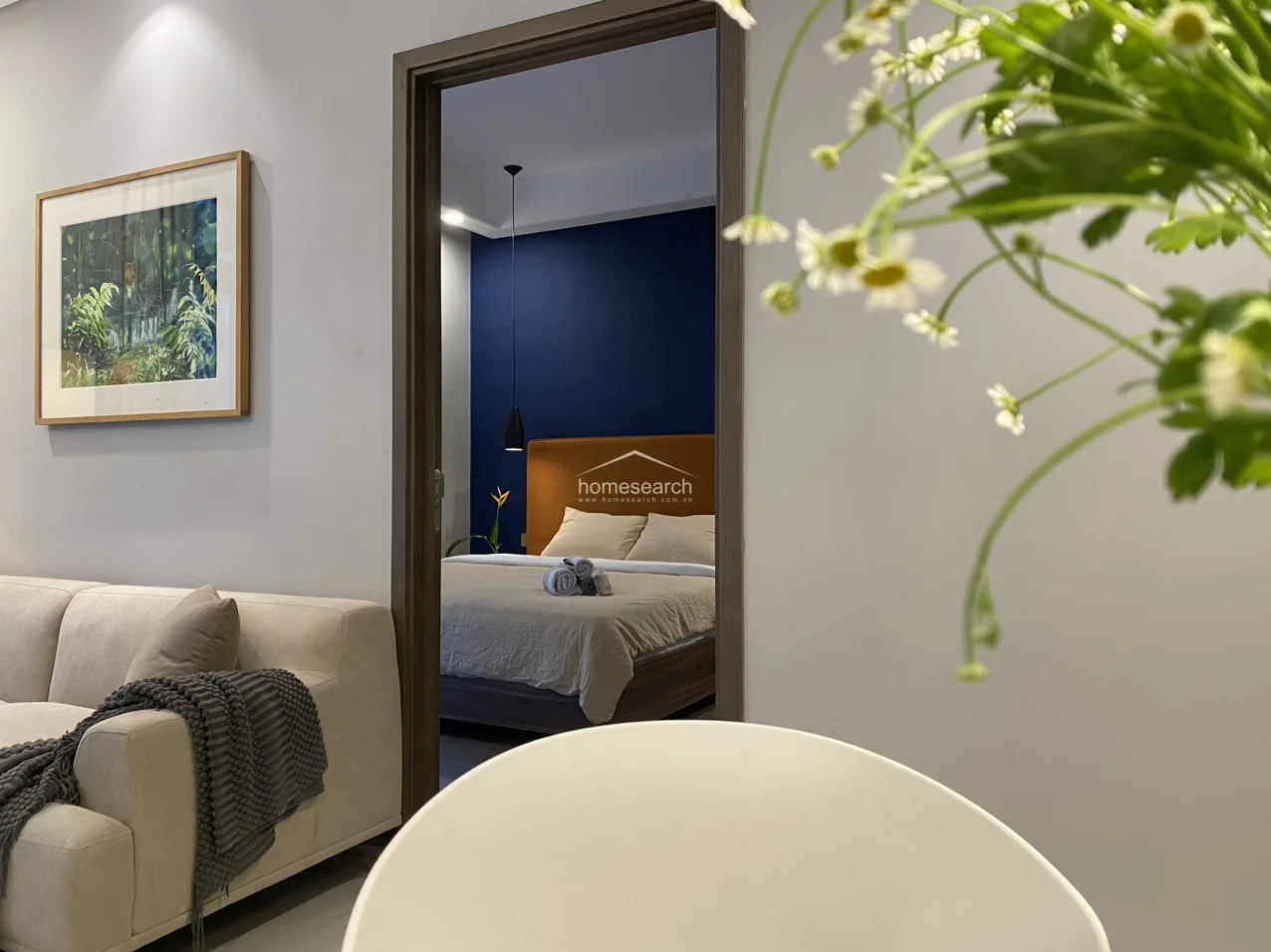This view from the living area into the master bedroom highlights the seamless transition between the communal and private spaces, emphasized by a clean, modern doorway. The interior design maintains a sophisticated color palette of neutral tones and deep blue accents, complemented by high-quality wood-look flooring and recessed ceiling lighting.