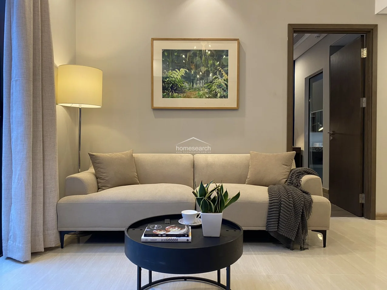 The seating area is centered around a comfortable beige sofa accented with a dark grey throw, perfectly complemented by a modern black circular coffee table. A floor lamp and framed nature-themed artwork add warmth, while the adjacent doorway offers a glimpse into a well-appointed bathroom.