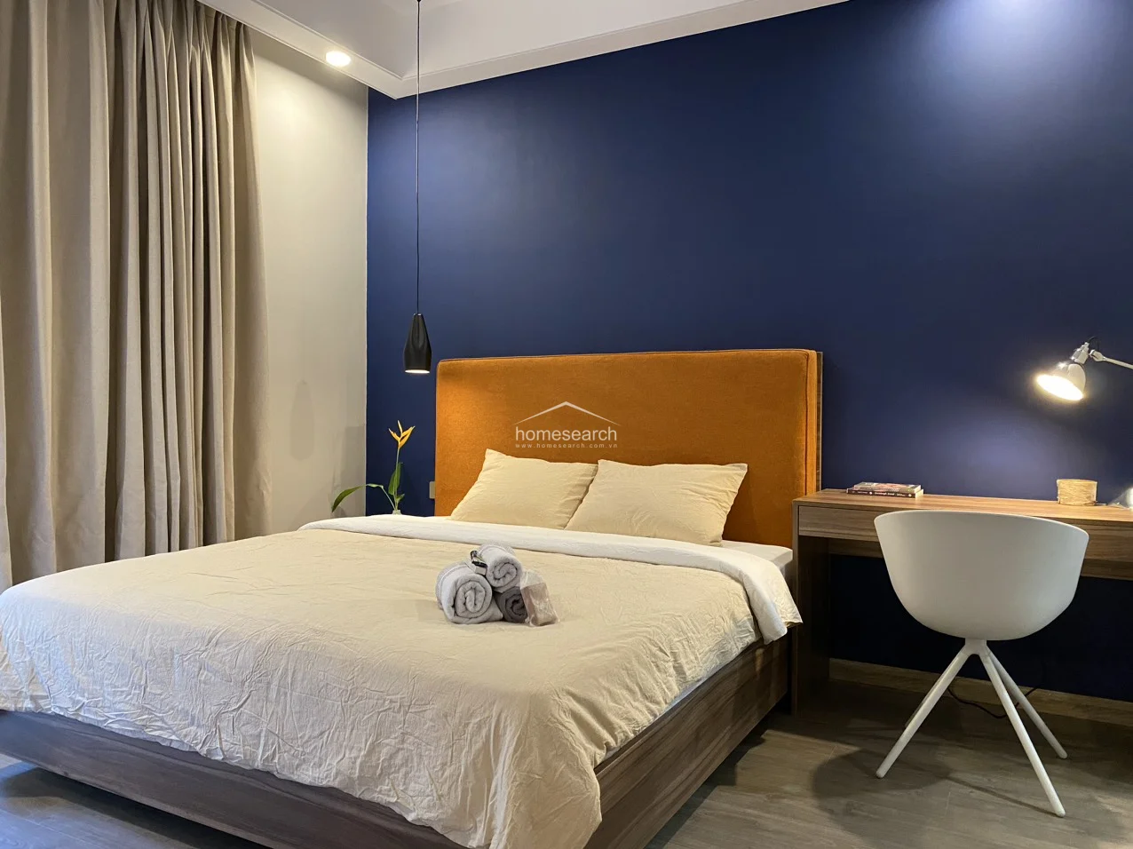 The master bedroom is thoughtfully furnished with a comfortable bed, a sleek wooden desk, and a minimalist pendant light that adds a touch of elegance. The space is well-maintained and features a cohesive aesthetic, utilizing a bold navy feature wall to anchor the room's modern, inviting atmosphere.