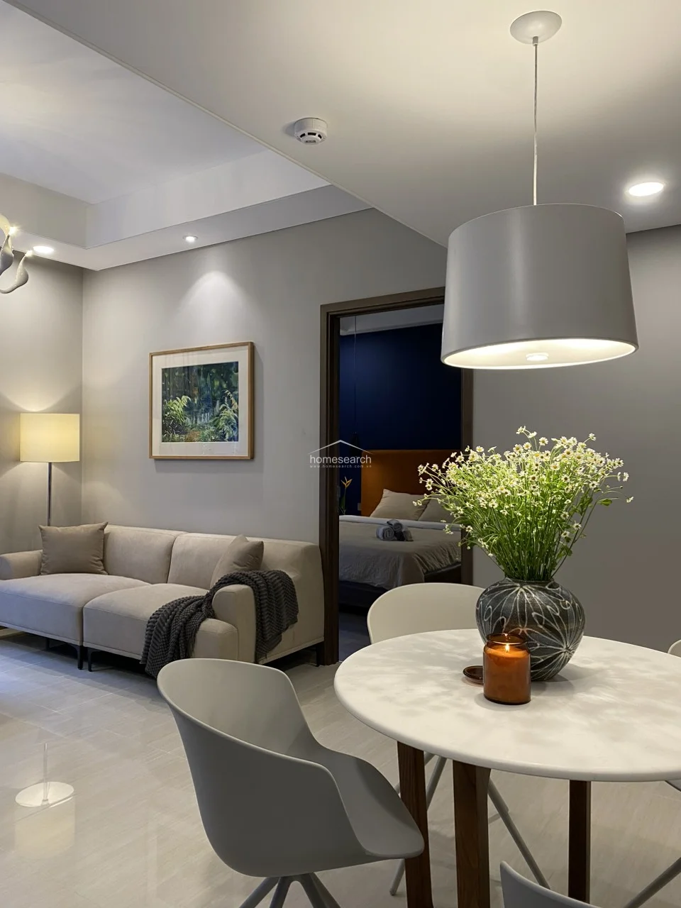 This open-concept living and dining area features a modern, neutral color palette with light grey walls and polished tile flooring. The space flows seamlessly into a bedroom visible through a doorway, while a stylish pendant light hangs over a circular dining table, creating an intimate atmosphere.