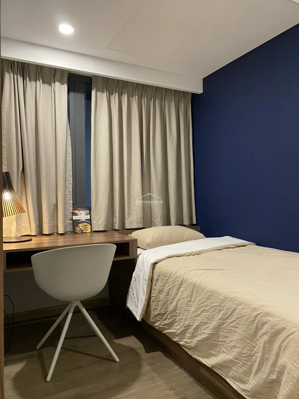 This bedroom features a contemporary design with a striking navy blue accent wall that contrasts beautifully with the warm, mustard-toned upholstered headboard. The room is equipped with a functional wooden desk, a modern white chair, and floor-to-ceiling beige curtains that provide privacy while allowing for soft, diffused natural light.