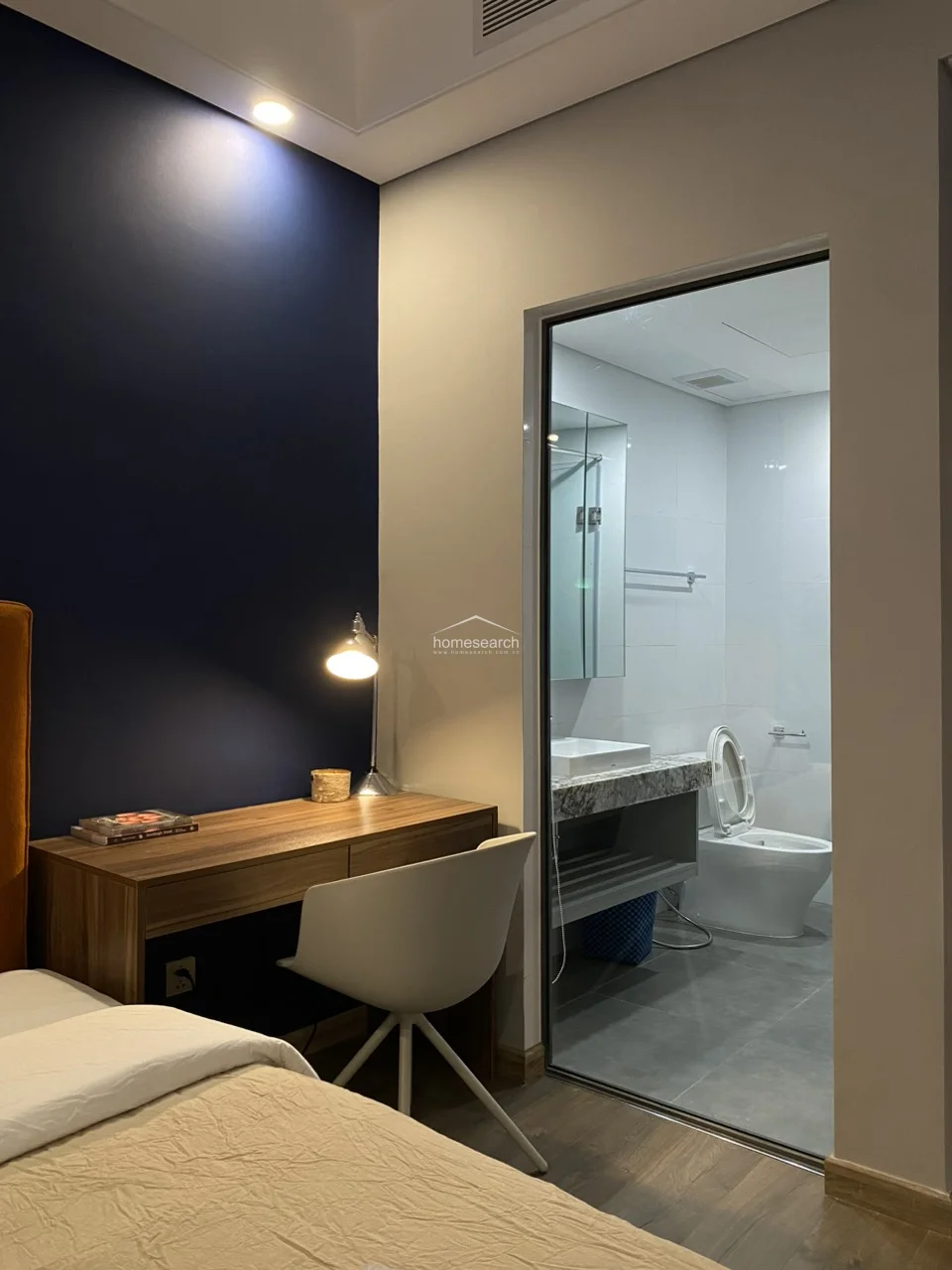 This image shows the master bedroom's direct connection to a modern ensuite bathroom, which is separated by a sleek glass partition to maximize the sense of space. The bathroom is finished with clean white wall tiles and a stone-topped vanity, reflecting a high level of maintenance and a contemporary, functional design.