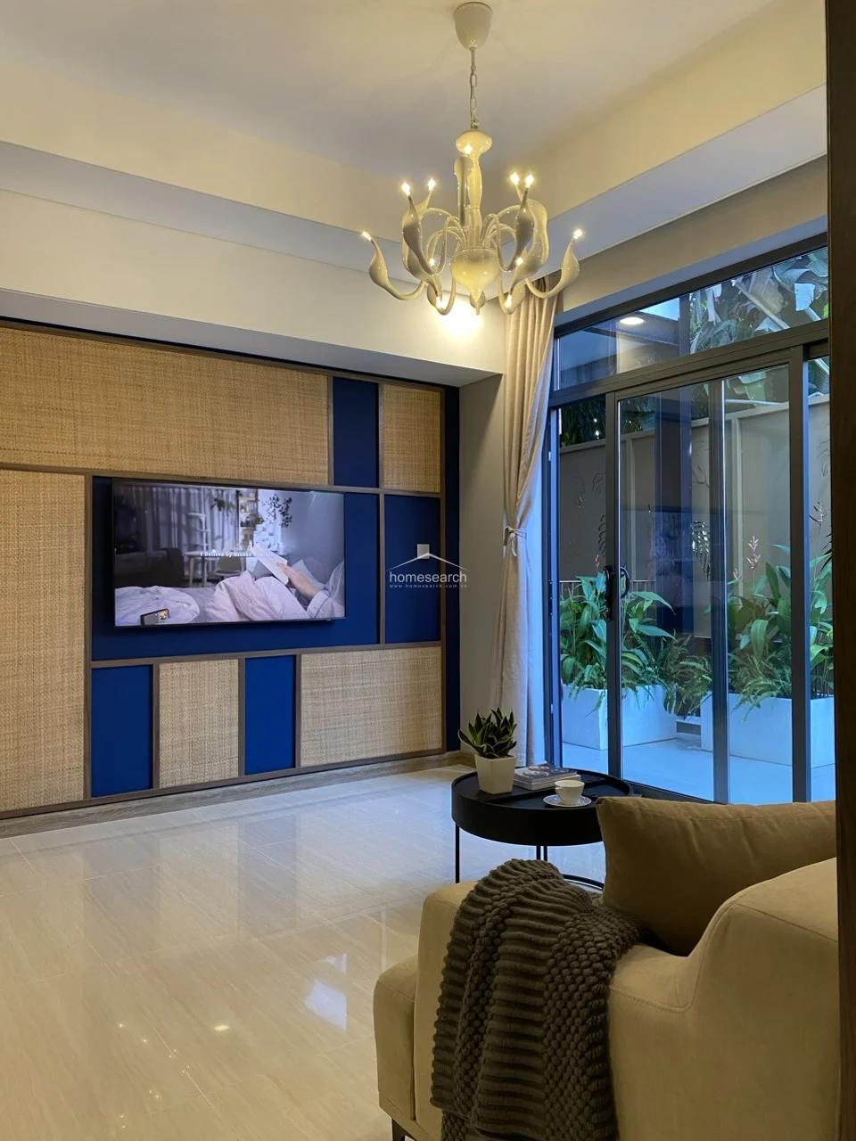 The living room is anchored by a feature wall combining deep blue accents with natural woven rattan panels, providing a sophisticated texture. Large sliding glass doors lead to an outdoor terrace filled with lush greenery, allowing ample natural light to brighten the room.