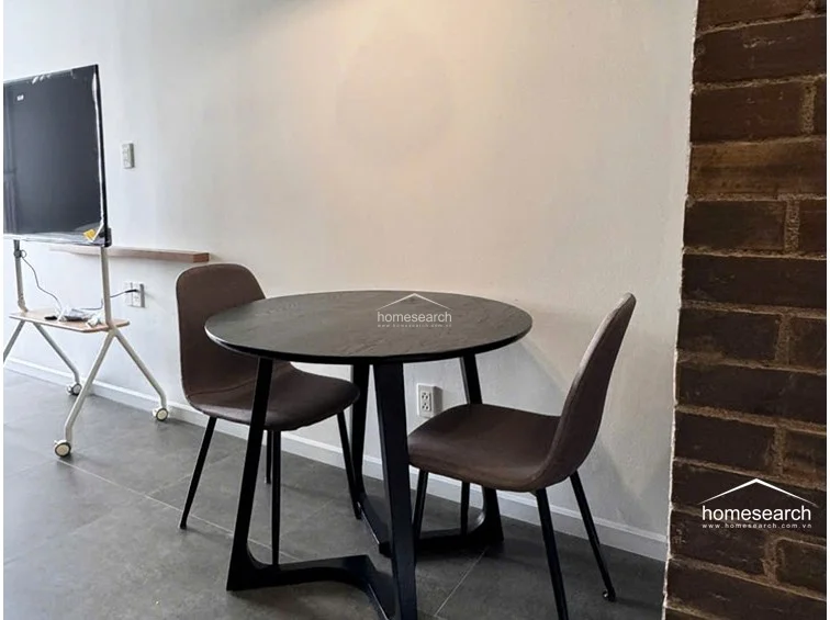 The dining area is compactly arranged with a round black table and two upholstered chairs, positioned against a textured brick wall that adds character to the room. A pendant light hangs above the table, while a nearby window provides a view into the bathroom, enhancing the sense of depth in the small space.