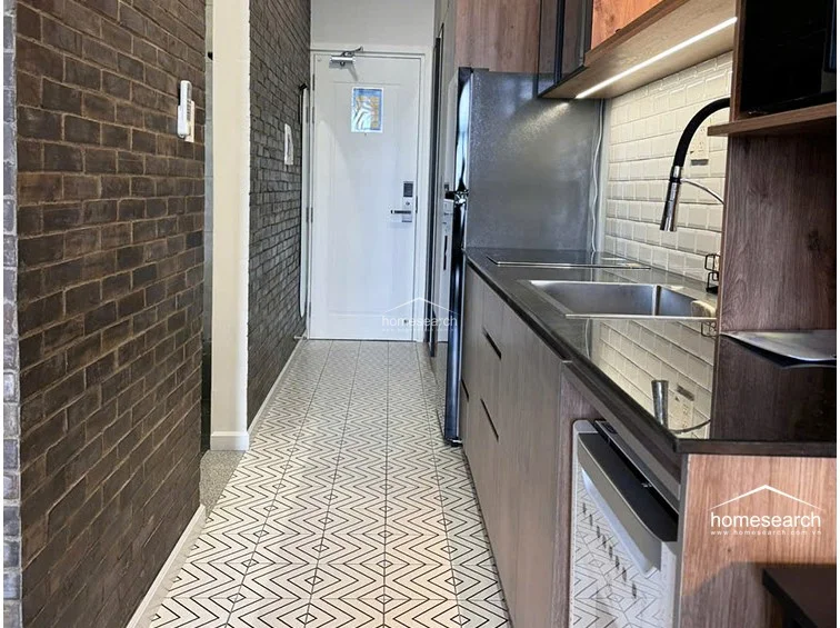 The kitchenette is a linear, galley-style design featuring wood-grain cabinetry, a stainless steel refrigerator, and a white subway tile backsplash. The floor transitions from the main living area's dark tiles to a decorative patterned tile, creating a distinct visual separation for the cooking zone.