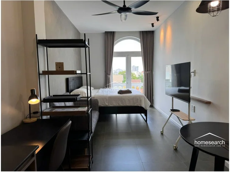 The sleeping and living area is anchored by a large bed and a grey sofa, illuminated by a modern black ceiling fan and recessed lighting. A tall, arched window with floor-to-ceiling curtains serves as the focal point, allowing for significant natural light and offering a view of the surrounding neighborhood.