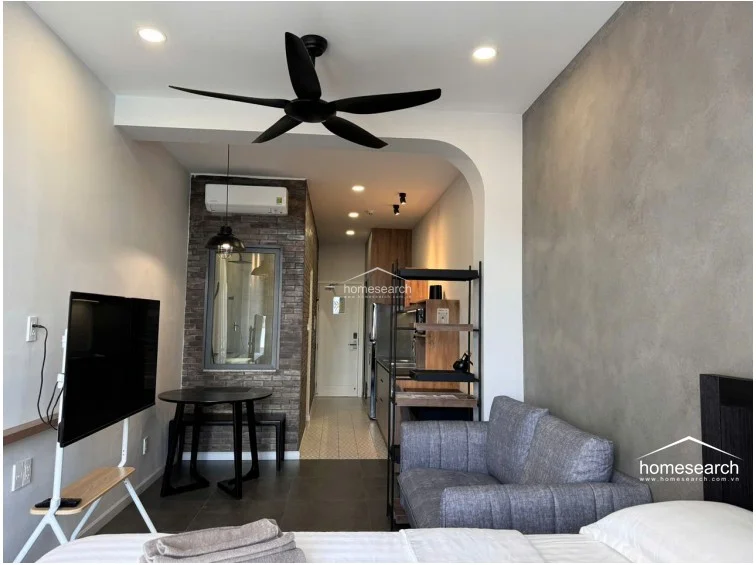 This view highlights the studio's smart use of vertical space, utilizing a black metal shelving unit as a room divider between the sleeping area and the kitchen. The room maintains a cohesive color palette of grey, black, and warm wood tones, creating a sophisticated and functional urban living environment.