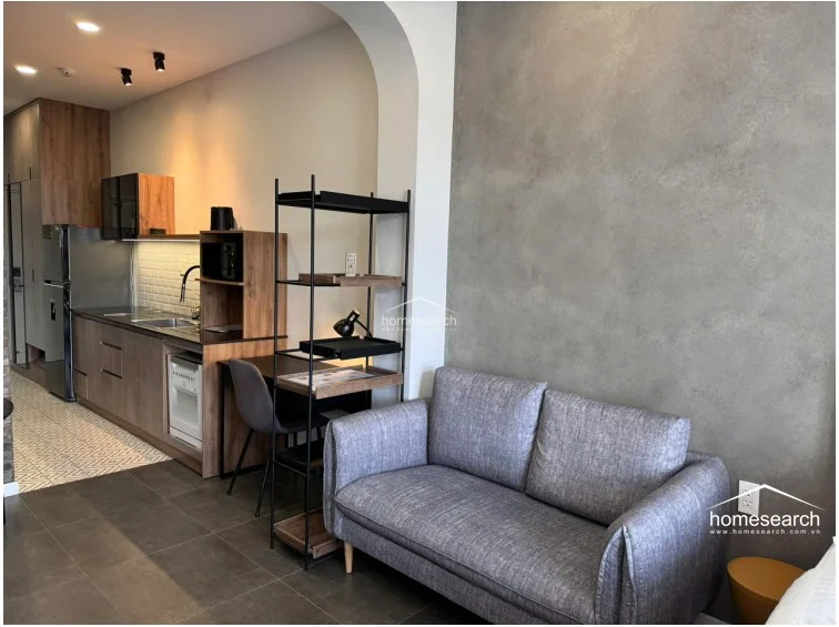 This open-concept living and kitchen area features a modern, industrial aesthetic with a textured grey accent wall and dark floor tiles. The space flows seamlessly from the kitchen into a cozy seating area, separated by a tall, open-frame shelving unit that serves as a functional room divider.