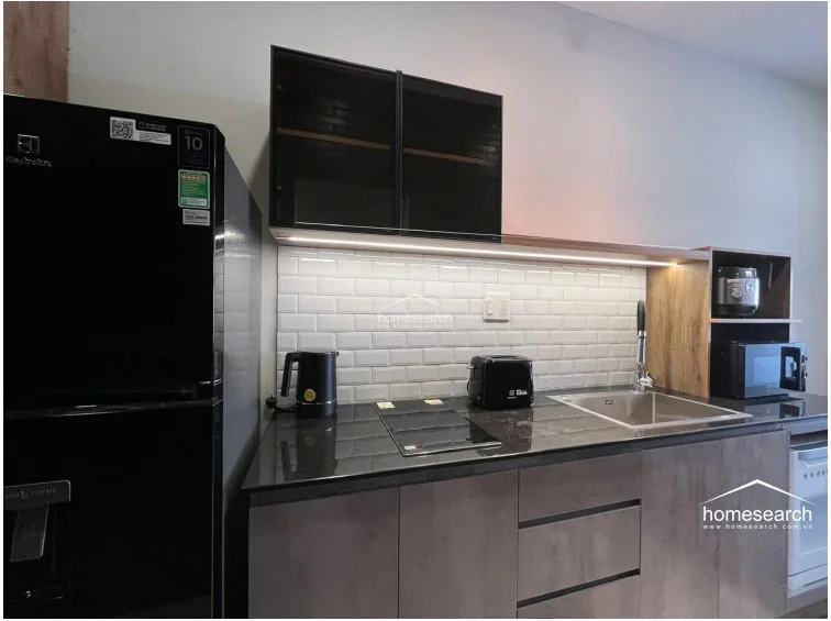 The compact kitchenette is highly efficient, featuring dark wood-toned cabinetry, a sleek black countertop, and a classic white subway tile backsplash. It is well-equipped with an Electrolux refrigerator, an integrated induction cooktop, and under-cabinet LED lighting for a clean, contemporary look.