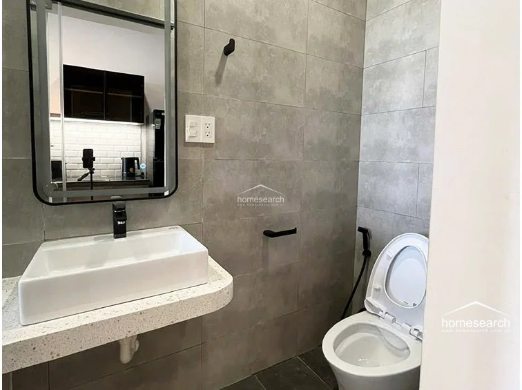 The bathroom maintains the apartment's modern theme with large-format grey wall tiles and a floating terrazzo-style vanity countertop. It includes a contemporary rectangular vessel sink, a sleek black-framed mirror, and matte black fixtures, all appearing to be in excellent, well-maintained condition.