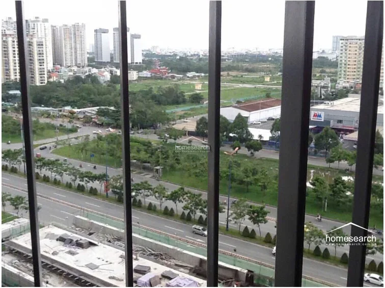 This image captures the elevated urban outlook from the balcony, which is protected by a sturdy metal railing. The view overlooks a mix of green spaces, local roads, and distant high-rise residential developments, suggesting a convenient city location. The balcony area is clean and well-maintained, offering a functional outdoor space for residents.