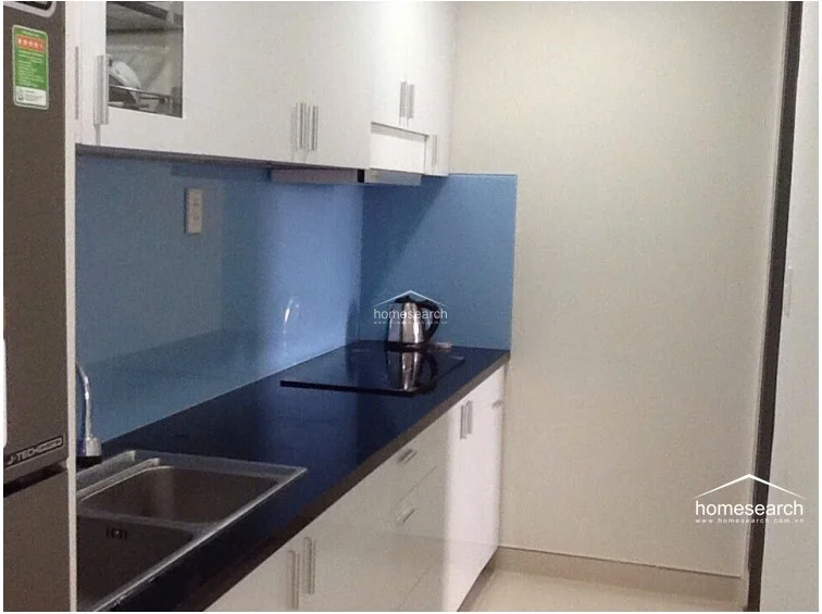 This galley-style kitchen is efficiently designed with white cabinetry, a contrasting dark countertop, and a vibrant blue glass backsplash that adds a pop of color. It is equipped with a built-in induction cooktop and a stainless steel sink, maintaining a clean and modern appearance. The space is well-lit and appears to be in excellent, move-in-ready condition.