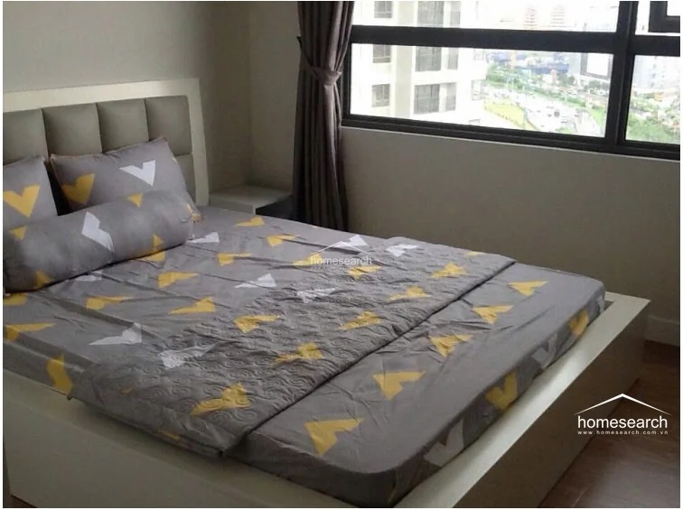 This bedroom focuses on a minimalist design, centered around a platform bed with a padded headboard and a geometric-patterned duvet. The room is brightened by a large window with dark curtains, and the wood-look flooring adds a touch of warmth to the otherwise neutral, well-kept interior.
