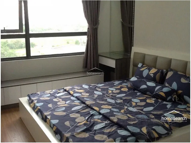 This bedroom features a contemporary design with a low-profile bed, a built-in low-level storage unit beneath a large window, and a matching vanity area. The room is finished with warm wood-tone flooring and neutral walls, complemented by floor-to-ceiling curtains that frame an expansive outdoor view.