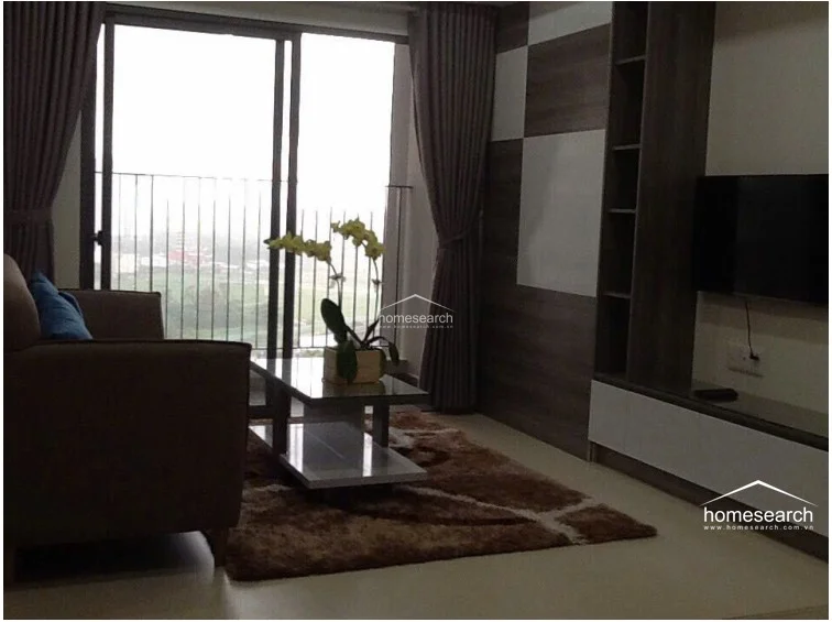 This living area features a modern, open-plan layout with large floor-to-ceiling glass doors that provide abundant natural light and access to a balcony with scenic views. The space is finished with neutral tile flooring and a sophisticated wood-paneled accent wall that incorporates built-in shelving and a wall-mounted TV console. The overall condition appears well-maintained, with contemporary furniture and elegant drapery enhancing the aesthetic.
