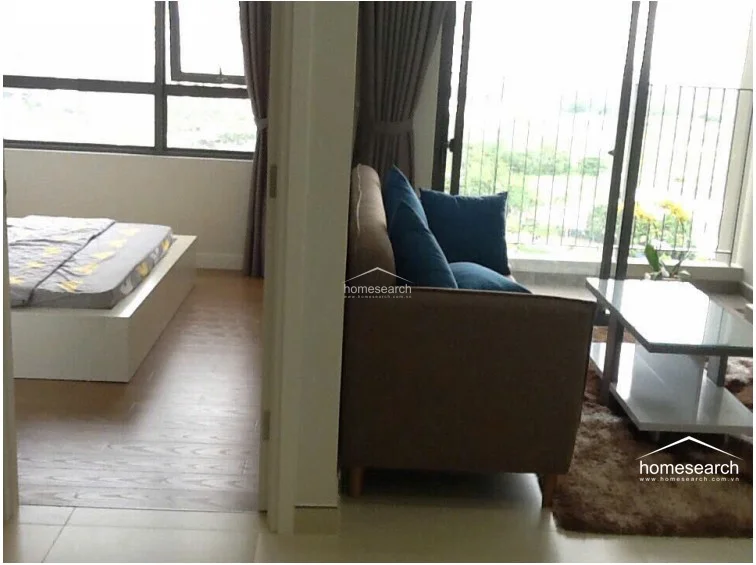 This image highlights the transition between the living room and the bedroom, showcasing a functional layout that maximizes space in a compact floor plan. The living area features tile flooring, while the bedroom transitions to warm wood-tone flooring, creating a distinct separation between the zones. Large windows in both areas ensure the apartment remains bright and connected to the outdoor environment.