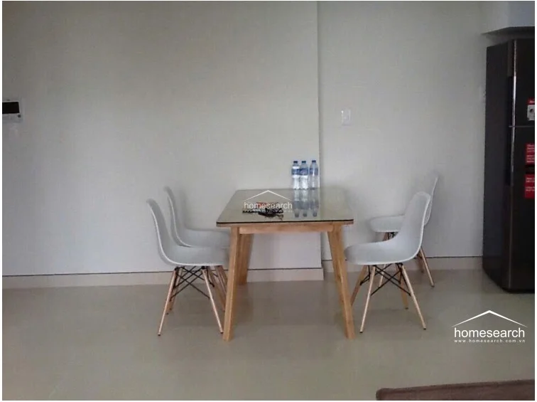 This dining area features a simple glass-topped table with four modern white Eames-style chairs, set against a neutral, minimalist wall. The space is finished with light-colored tile flooring and includes a tall, dark-toned refrigerator nearby, suggesting an open-plan layout that flows into the kitchen.