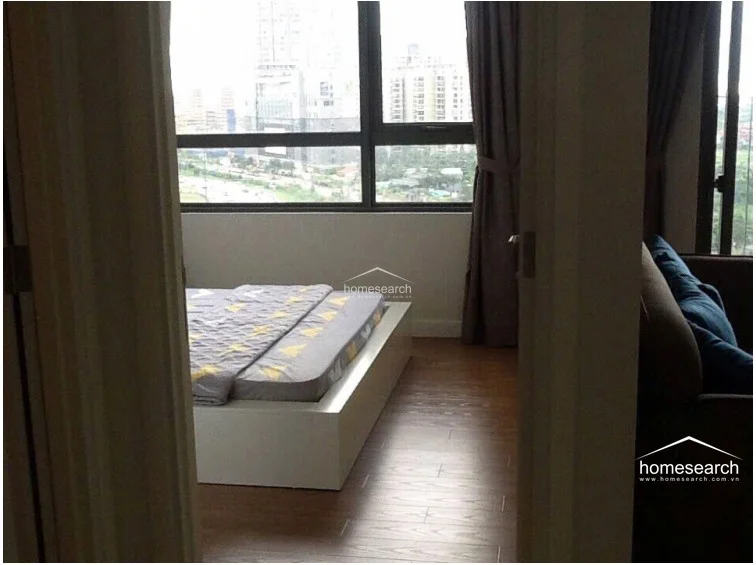 This bedroom offers a cozy, functional layout with a low-profile platform bed and wood-look flooring that provides a warm contrast to the neutral walls. A large window with full-length curtains allows for ample natural light and provides an elevated urban view, while the room maintains a clean, well-maintained aesthetic.