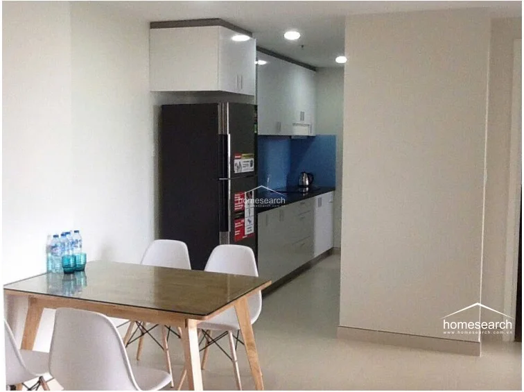 This dining and kitchen area features a clean, minimalist design with a glass-topped dining table and white modern chairs. The kitchen is equipped with high-gloss white cabinetry, a dark countertop, and a blue glass backsplash, complemented by a full-sized refrigerator. The space is bright and functional, with recessed ceiling lighting and light-colored tile flooring that enhances the open, airy feel of the apartment.