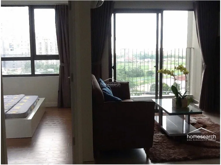 This view highlights the open-concept flow between the living room and the adjacent bedroom, which is separated by a doorway. The bedroom features warm wood-look flooring, while the living area maintains a consistent neutral aesthetic, both benefiting from ample natural light through large windows and balcony doors. The overall layout feels cohesive and well-organized, with heavy curtains providing privacy and light control.