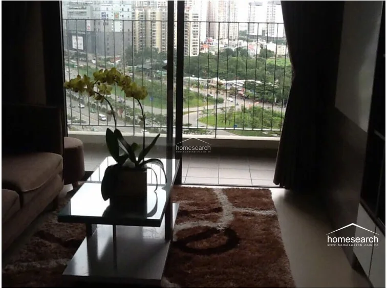 This living room area opens directly onto a balcony through large sliding glass doors, providing ample natural light and an elevated urban view. The space is furnished with a comfortable sofa, a glass-topped coffee table, and a plush area rug, all set against a neutral color palette that emphasizes a relaxed, well-maintained atmosphere.