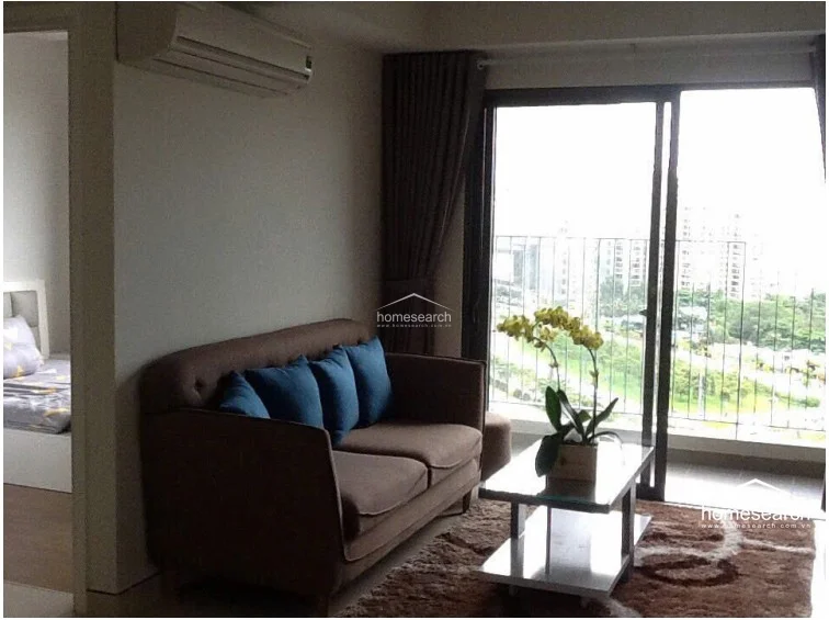 This perspective emphasizes the living room's connection to the bedroom and the balcony, showcasing a wall-mounted AC unit for climate control. The room is furnished with a comfortable brown sofa, a glass-topped coffee table, and a plush rug, creating a cozy yet modern atmosphere. The neutral walls and dark curtains provide a balanced contrast, reflecting a high standard of interior maintenance.