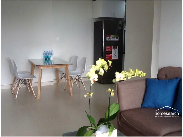 The dining area is seamlessly integrated into the open-concept living space, featuring a compact glass-top table with four modern, minimalist chairs. A full-sized refrigerator is positioned nearby, suggesting a functional flow between the kitchen and the social areas. The neutral wall colors and consistent flooring create a cohesive and airy atmosphere throughout the room.