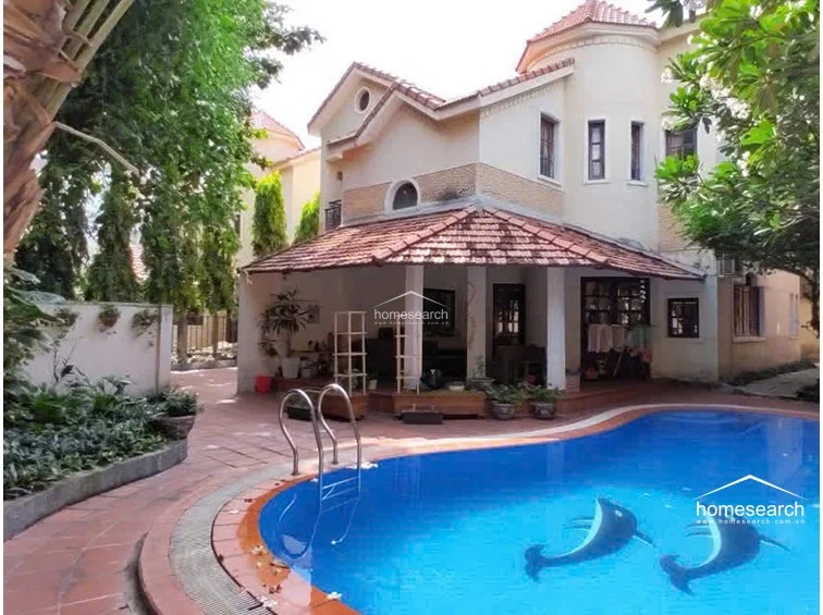This exterior view showcases a multi-story villa featuring a classic terracotta-tiled roof and a prominent turret-style architectural element. The property includes a private, kidney-shaped swimming pool with decorative dolphin motifs and a shaded, tiled patio area that provides a seamless transition between indoor and outdoor living spaces.