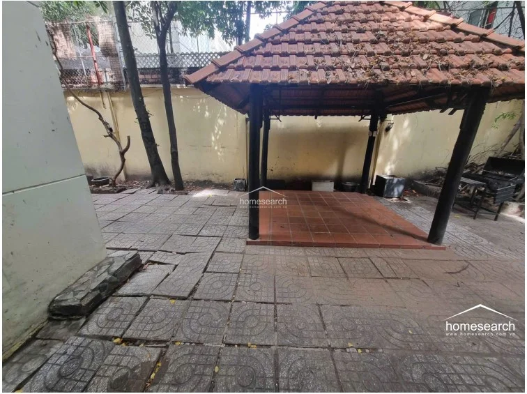 This image shows a garden patio area anchored by a traditional wooden gazebo with a tiled roof, providing a shaded outdoor seating or dining spot. The surrounding ground is paved with patterned concrete tiles, and the area is bordered by high walls and mature trees, offering a secluded, quiet outdoor retreat.