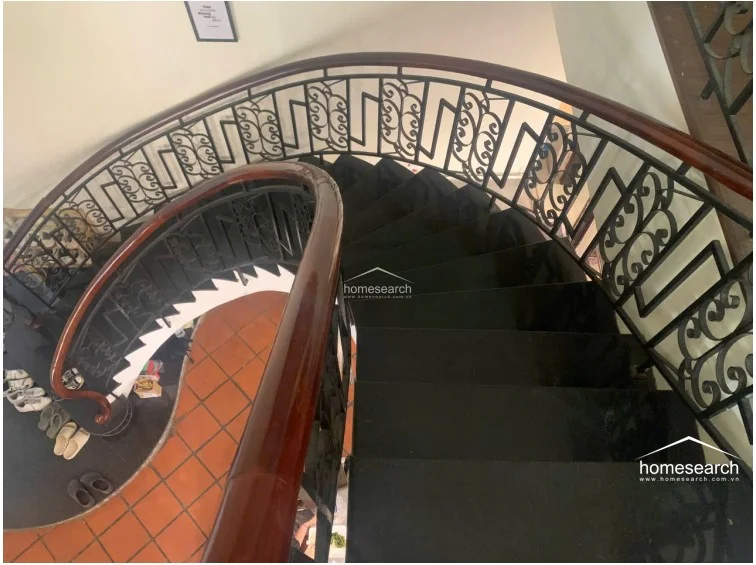 This elegant spiral staircase serves as a focal point, featuring dark stone treads and a decorative black wrought-iron railing topped with a polished wooden handrail. The surrounding area is floored with traditional terracotta tiles, and the architectural design emphasizes a classic, high-end aesthetic with a smooth, curved flow.