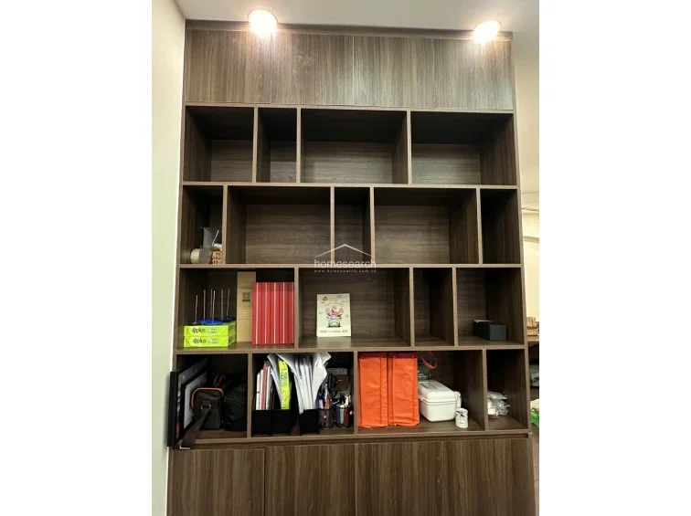 This floor-to-ceiling wooden storage unit features a modern, asymmetrical grid design with open shelving and closed cabinets at the base. The dark wood finish provides a sophisticated contrast to the neutral walls, and the integrated overhead spotlights ensure the unit is well-lit for display or organizational purposes.