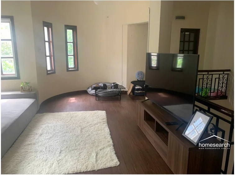 This living area features a unique curved wall design with multiple vertical windows that provide natural light and a view of the outdoors. The space is finished with dark wood-look flooring, cream-colored walls, and includes a modern media console and a plush white area rug, creating a cozy yet functional layout that connects to a staircase landing.
