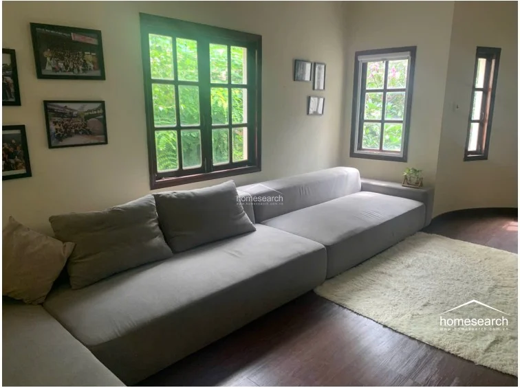 This cozy living room is furnished with a large grey sectional sofa and a soft area rug, set against warm-toned walls. Multiple windows with dark wood frames allow for natural light and provide a pleasant view of the surrounding greenery, creating a comfortable and inviting atmosphere.