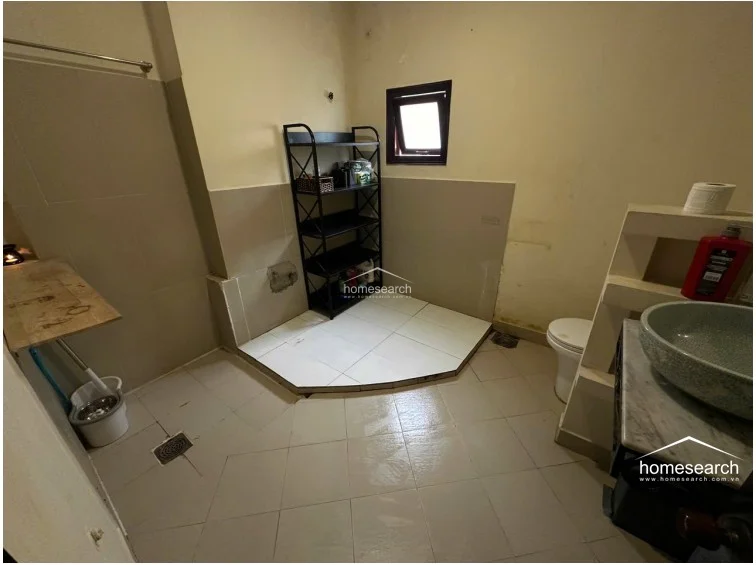 This bathroom features light-colored floor and wall tiles with a raised, tiled platform area for storage or a shower setup. It includes a vessel sink on a marble-topped vanity and a small window for ventilation, though the overall condition appears dated and in need of refreshing.