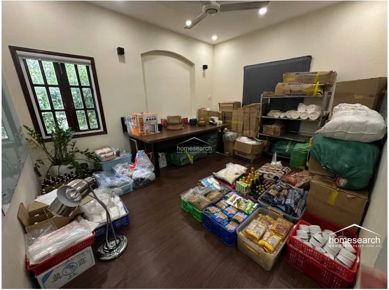 This room is currently utilized as a storage or utility space, filled with various boxes, crates, and shelving units containing household goods and supplies. It features dark wood flooring, cream walls, and a ceiling fan, with a window providing natural light and a view of the exterior greenery.