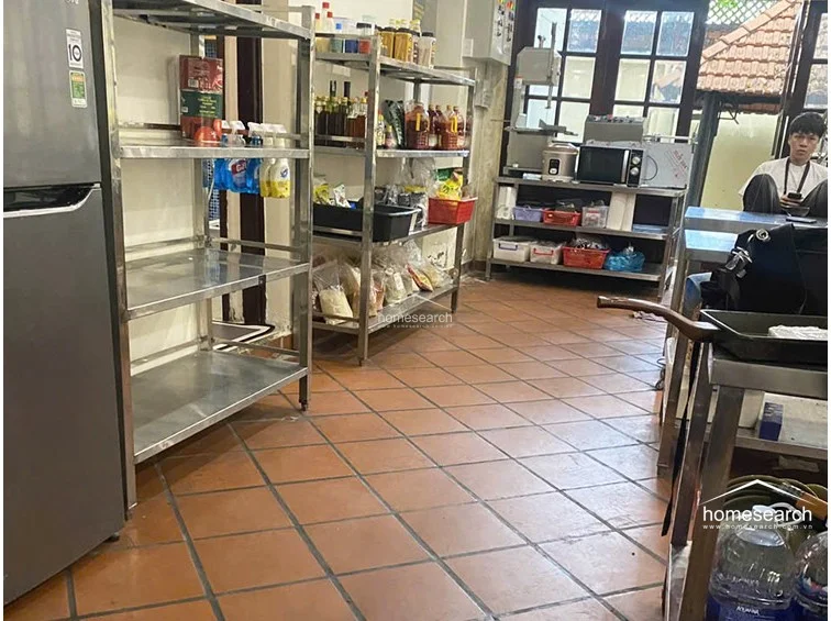 This functional kitchen or pantry area is equipped with industrial-style stainless steel shelving units and a refrigerator, indicating a practical, high-utility layout. The floor is covered in durable terracotta tiles, and the space connects to the rest of the home via a dark-framed glass door, maintaining a utilitarian and well-organized condition.