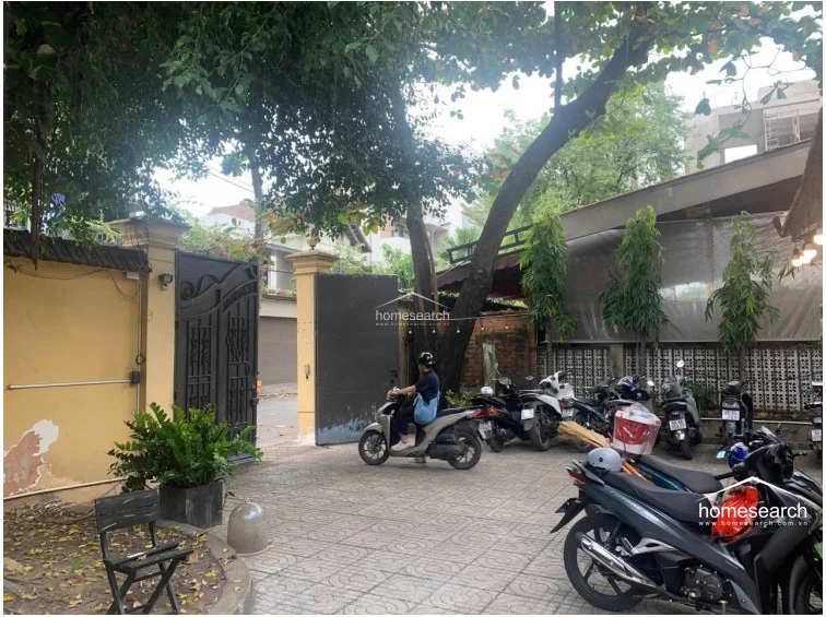 This outdoor courtyard serves as a functional parking area for motorbikes, enclosed by yellow perimeter walls and a sturdy metal gate. The space features paved tiling and mature trees that provide natural shade, though the overall condition shows signs of wear, including peeling paint on the exterior walls.