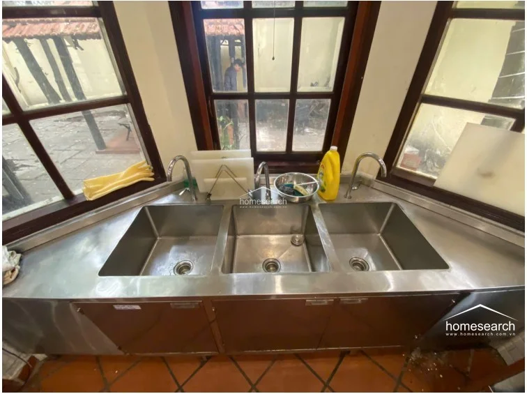 This kitchen area features a unique, multi-basin stainless steel sink unit set against a bay window configuration, allowing for ample natural light. The flooring is finished in traditional terracotta-style tiles, and the dark wood window frames provide a rustic contrast to the industrial-style metal cabinetry below the sink.