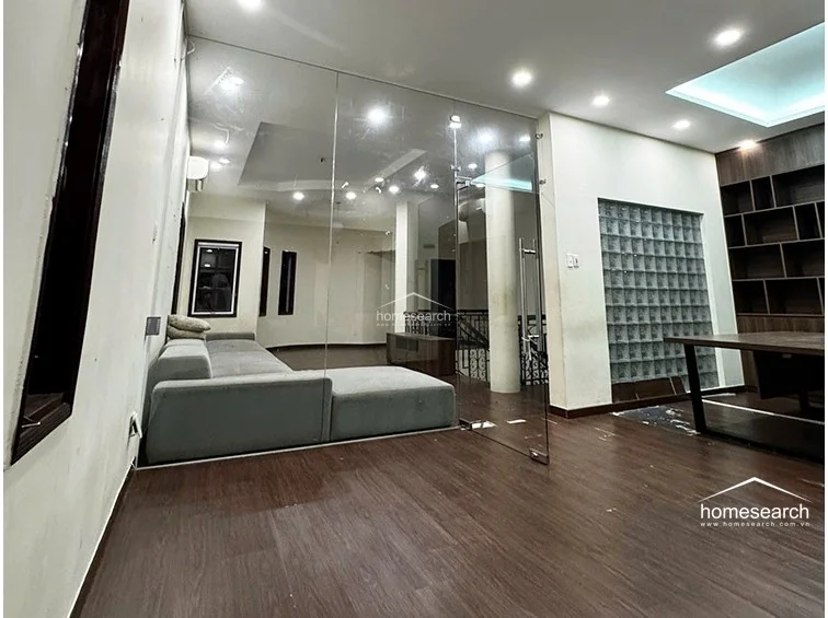 This multi-functional space is divided by a floor-to-ceiling glass partition, allowing for a seamless visual flow between the living area and an office or study zone. The room includes a built-in wooden shelving unit, a glass block feature wall, and recessed ceiling lighting, highlighting a contemporary and organized design.