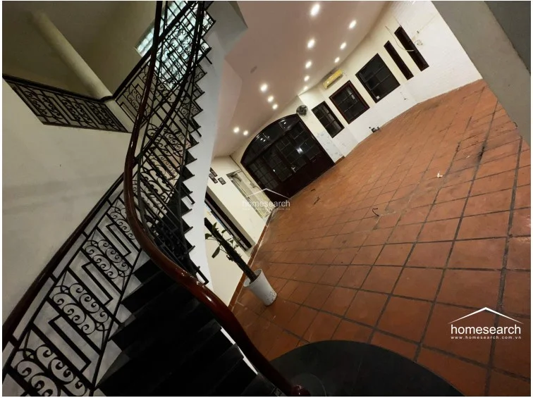 This spacious, open-plan room features a striking spiral staircase with a decorative wrought-iron railing and polished wooden handrail. The area is finished with terracotta tile flooring, recessed ceiling lighting, and a large arched glass entryway, presenting an expansive layout that is currently unfurnished and well-maintained.