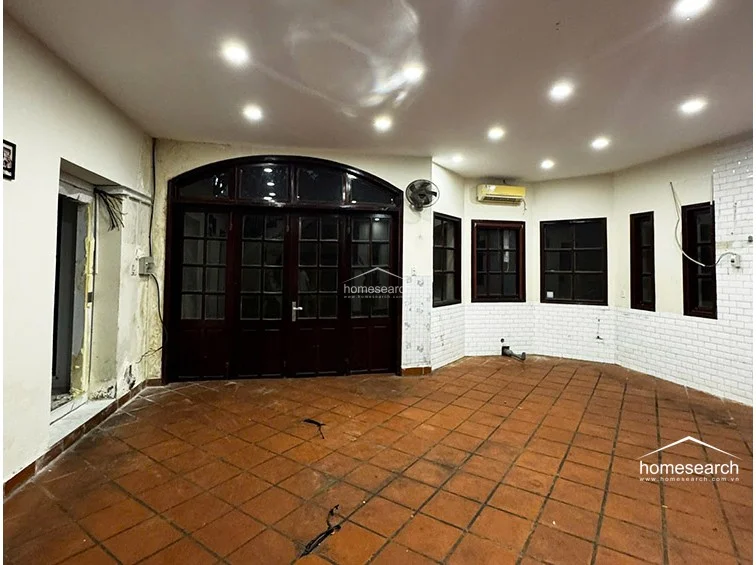 This spacious living area features terracotta-style floor tiles and white walls partially accented with white subway-style tiling. The room is illuminated by numerous recessed ceiling lights and includes a wall-mounted air conditioning unit, a ceiling fan, and a large, dark-wood arched glass door that provides natural light and exterior access.