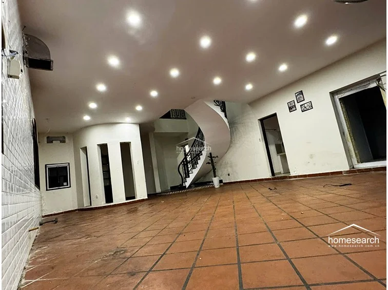 This view highlights the open-plan layout of the living space, which connects to other areas via a prominent, curved white staircase with a black metal railing. The room maintains a consistent aesthetic with terracotta flooring and recessed lighting, though some wall areas show signs of wear and require maintenance.